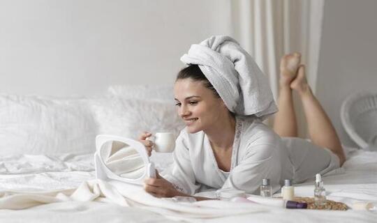 Beautiful brunette woman with a white robe and a towel wrapped around her head, looking in the mirror drinking a cup of coffee ready to give herself a self-care treatment at home.