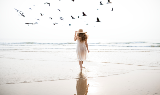 A young lady walking by the seashore in a white dress, one hand on her sun hat, relaxing and alleviating her stress.