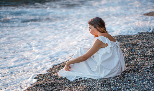 Beautiful young woman alone sitting on a seashore in a white dress thinking about solutions to her problems that are rolling in like a flood.