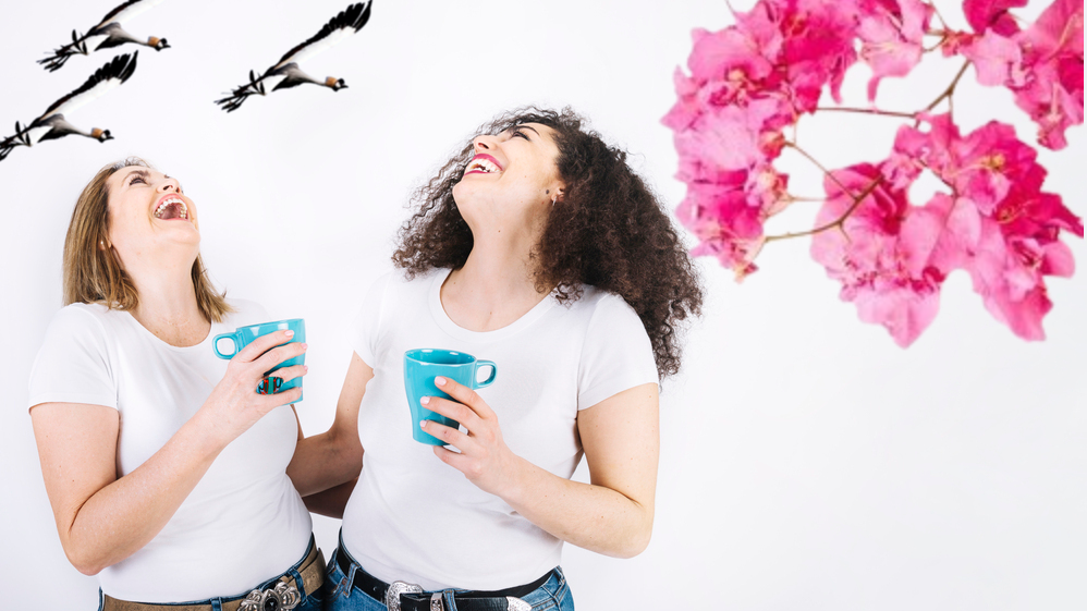 Two ladies wearing white Tee shirts laughing hardy while each holds a coffee cup in their hands.